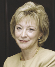 A woman with chin length blonde hair and bangs wears a beige top and gold necklace.