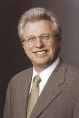 A man with white hear wears glasses, a light brown suit jacket, white button down, and gold tie.