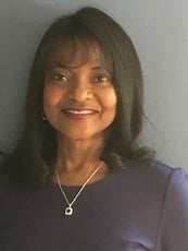 A woman with a shoulder length black hair and bangs wears a purple top