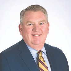 A man wears a dark blue suit jacket, a light blue button down, and a red, blue, and yellow striped tie.