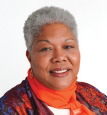 A woman with grey curly hair wears a bright orange scarf and a dark red, blue, and orange cardigan.