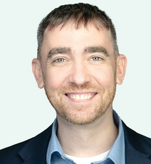 A man with a scruffy beard wears a dark blue suit jacket, a light blue button down, and a white undershirt.