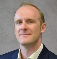 A man with blonde hair wears a dark blues suit jacket and white button down.