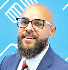 A bald man with a brown beard wears large black glasses, a dark blue suit jacket, a white button down, and a red, yellow, grey, and black patterned tie.