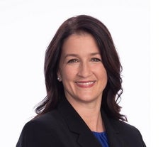 A woman with long dark brown hair wears a black suit jacket and blue top.