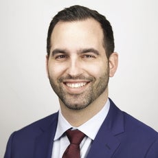 A man with short brown hair wears a dark blue suit jacket, a white button down, and a dark red tie.