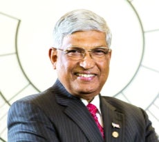 A man with white and grey hair wears a black pinstriped suit, a white button down, and a pink and red patterned tie.