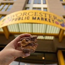 A hand holds up a chocolate and peanut butter treat in front of a big yellow sign reading "Worcester Public Market".