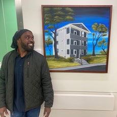 A man wearing a dark green jacket, dark blue shirt, and jeans stands against a wall looking at a painting of a grey triple-decker house in front of a blue sky with trees on either side of it.