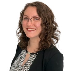 A woman with medium-length curly brown hair wears glasses, a black suit jacket, and a black, pink, and light green patterned top.