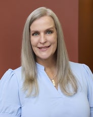 A woman with long, straight grey hair wears a baby blue v-neck top.