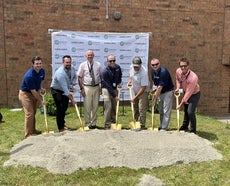 Men stand with shovels next to a pile of dirt on a plot of grass in front of a brick building.