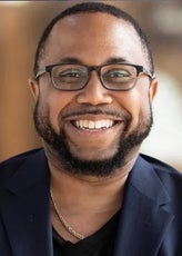 A man with short black hair and a beard wears black glasses, a dark blue suit jacket, a black top, and a gold chain around his neck.