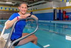 A young woman with light brown hair pulled back sits at the edge of a pool with her arms on a railing wearing a light blue headband, a blue top, and black shorts.