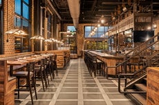 Inside a restaurant with high ceilings and a checkered floor with wooden tables and high chairs on the left and a bar on the right