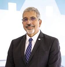 A man with grey hair and goatee wears thin-wired glasses, a black suit jacket, a white button down, and a blue striped tie.