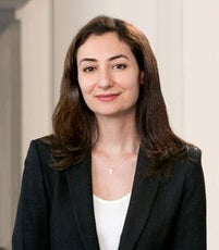 A woman with long dark brown hair wears a black suit jacket and a white top.