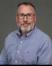 A man with grey and white hair and beard wears black glasses and a blue and white button down.