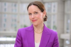 A woman with light brown hair tied back in a bun wears pink lipstick, an electric blue suit jacket, a white v-neck top, gold necklace, and pearl earrings.