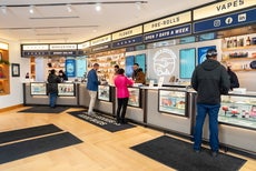 Inside a cannabis dispensary with four cashiers standing behind a counter with windows featuring products and four customers checking out