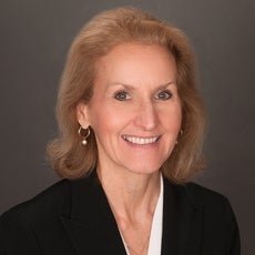 A woman with shoulder length blonde hair wears a black blazer, white top, and pearl necklace.