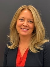 A woman with long blonde hair wears a black suit jacket and red top.