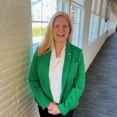 A woman with long blonde hair wears a green blazer and white top while standing in a hallway next to a white brick wall with windows.