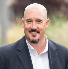 A man with a dark brown goatee wears a dark blue suit jacket and white button down while standing outside.