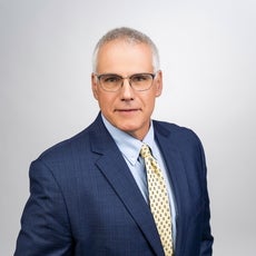 A man with white hair wears black glasses, a dark blue plaid suit jacket, a light blue button down, and a yellow patterned tie.