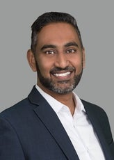 A man with black and grey hair and beard wears a dark blue suit jacket and white button down.