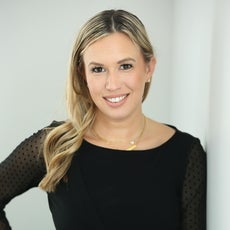 A woman with long blonde hair leads against a white wall with one hand on her hip while wearing a black top.