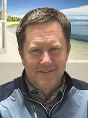 A man with short brown hair smiles in front of an image of a beach while wearing a dark and light blue zip-up sweater.