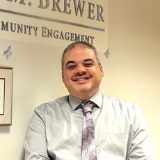 A man with grey hair and a goatee wears a grey button down and a purple and grey paisley tie.