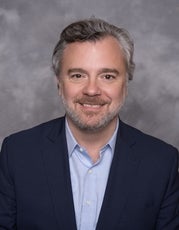 A man with short wavy grey and white hair smiles for the camera wearing a dark blue suit jacket and light blue button down.