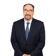 A man with brown and grey hair and goatee wears black glasses and stand with his arms together in front of him while wearing a dark blue suit.