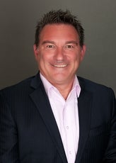 A headshot of a man with spiked up dark brown hair wearing a dark blue pinstriped suit jacket and light pink button down.