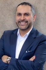 A man with black and grey hair and beard wears a dark blue suit jacket and white button down.