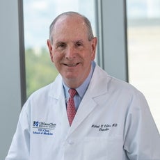 A man with grey hair wears a white lab coat, a light blue button down, and a red patterned tie.