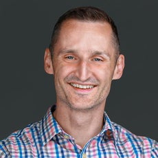 A man with light brown hair wears a white, blue, pink, and grey plaid shirt.