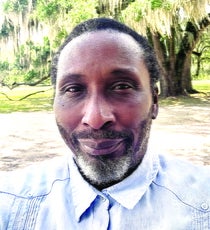 A man with short black and grey hair and beard wears a light blue button up while standing outside in front of lush trees.