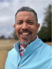 A man with short curly black and grey hair and a beard wears a light blue zip up sweater while standing outside.