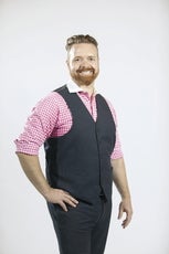 A man with a red beard smiles at the camera wearing a pink checkered shirt and navy vest.