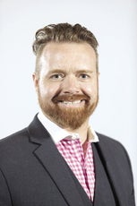 A man with red hair and a beard smiles for the camera wearing a grey suit jacket and white and pink plaid button down.