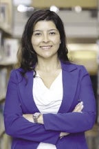 A woman with long brown hair wears a blue suit jacket and white blouse with her arms crossed.