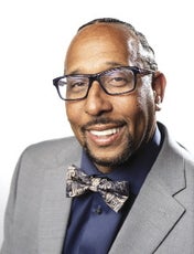 A man wears black glasses, a grey suit jacket, dark blue button up, and patterned bow tie.