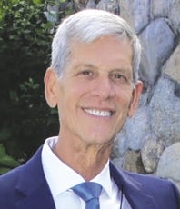 A man wears a dark blue suit jacket, white button down, and blue tie in front of a stone wall.