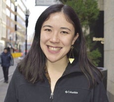 A woman with dark hair wears a black zip up jacket and dangly earrings.