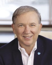 A headshot of a man with grey hair wearing a dark blue suit jacket and white button down