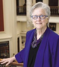 A woman with short grey hair wears purple glasses, a purple jacket, black top, and beaded green and purple necklace with her hand on a railing