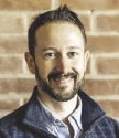 A man with a beard stands in front of a brick wall